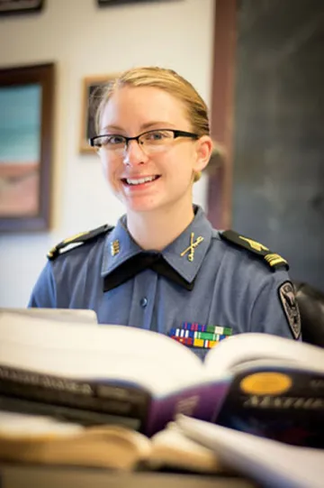A young woman in formal military shirt smiles as photographed behind an open book at a desk. Photo of Rebecca Sweem '16, NUCC Regimental Commander 2015-2106.