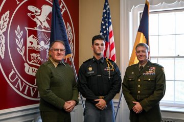 Three people in uniform stand in front of a large university seal and two flags.