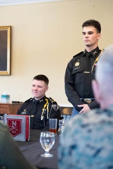 A person in a dark uniform stands beside another seated at a table set with drinks, dishes, and a red Norwich napkin holder.