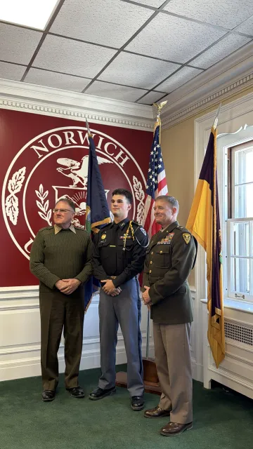Three people stand in front of a Norwich University seal and two flags in a room with white trim and green carpet.