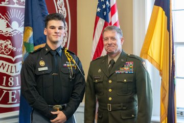 Two people in military uniforms stand indoors in front of U.S. and colored flags near a wall with a university seal.