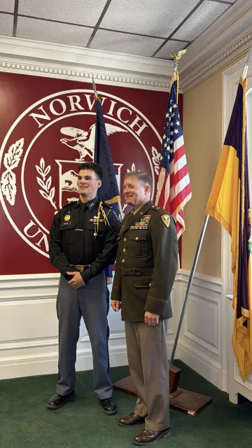  Two people in military uniforms stand in front of a Norwich University seal and three flags.
