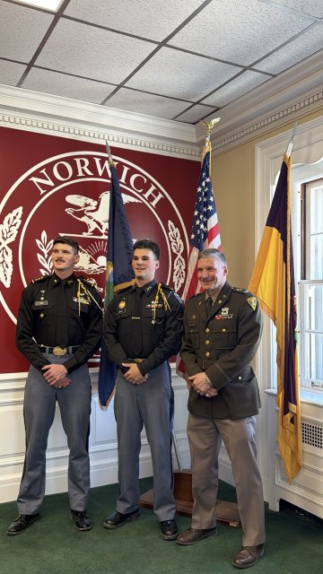 Three people in military uniforms stand in front of the Norwich University seal and three flags in a room with green carpet.