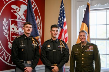 Three people in military uniforms stand indoors in front of a large Norwich University seal and three flags.