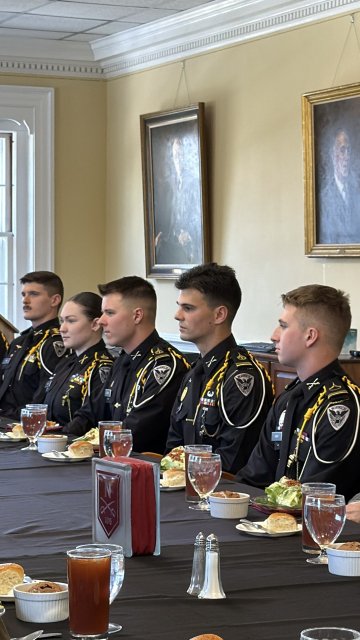 People in formal uniforms sit at a long table set with plates of food, drinks, and silverware in a dining room with framed portraits on the wall.