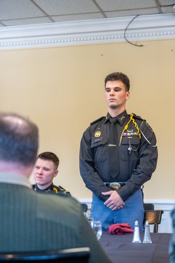 A person in a dark uniform stands at a dining table with plates, glasses, and salt and pepper shakers.