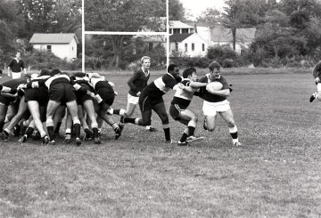 Black and white photo of rugby action, with one rugger with ball moving away from two opponents and the remainder of the players in a scrum. Rugby game, Norwich University vs. West Point, 1976.