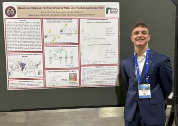 Student stands beside a research poster on removing uranium from drinking water with a peptide filter.