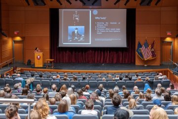 Student presents to an audience in an auditorium with a slide projected on stage.