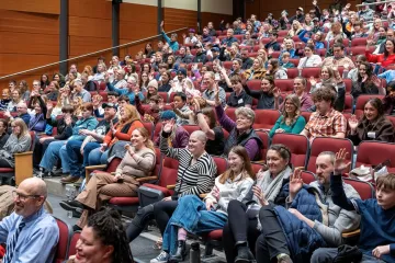 Audience members raise their hands while seated in an auditorium.