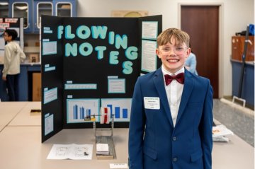 Student in a blue suit stands beside a Flowing Notes science fair display.