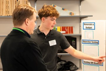 Student points to a science fair display while speaking with an adult.