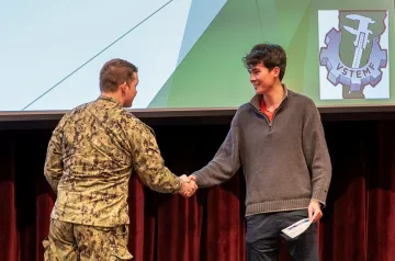 Person in uniform shakes hands with a student holding a paper on a stage.