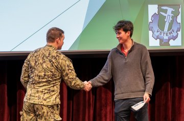 Person in uniform shakes hands with a student holding a paper on a stage.