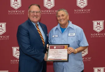 A man and staff recipient Mary O'Callaghan hold a Norwich University award certificate in front of a branded backdrop.