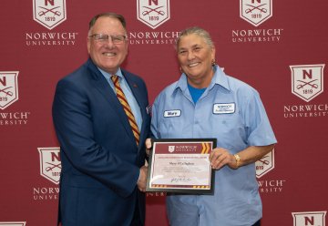 A man and staff recipient Mary O'Callaghan hold a Norwich University award certificate in front of a branded backdrop.
