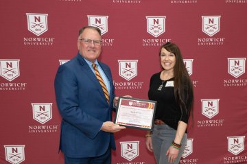 A man presents a Norwich University award certificate to a staff recipient in front of a branded backdrop.