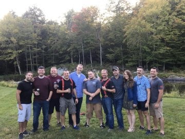 A group of individuals, some holding beverages, stand outside in front of a pond and a stand of trees.
