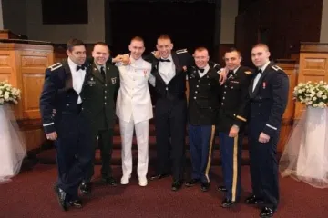Seven individuals in formal attire stand indoors before a raised platform, with floral arrangements on either side.