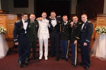 Seven individuals in formal attire stand indoors before a raised platform, with floral arrangements on either side.