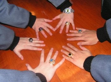 Six hands are shown together on a table in a circular pattern, each hand bearing a jeweled class ring.
