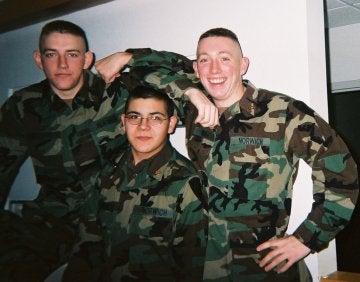 Three cadets in military fatigues stand together, two with arms over head of third.