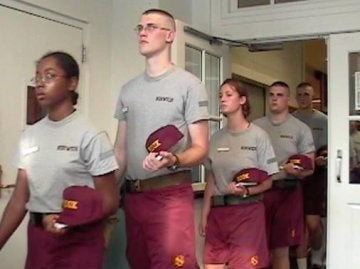 A line of individuals in grey t-shirt, red shorts, holding red caps, walking into a room through a doorway.
