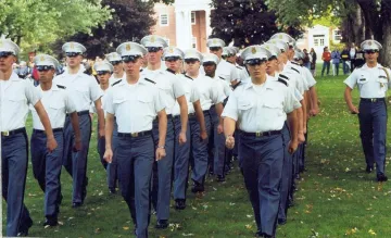 Group of military students in white cap, white shirt and grey pants march in formation on a grassy lawn.