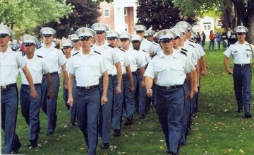 Group of military students in white cap, white shirt and grey pants march in formation on a grassy lawn.