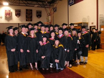 Group of graduates in cap and gown stand or kneel together on a basketball court.