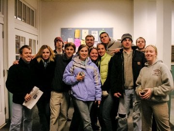 A large group of individuals, most in jackets, stand in room before a bulletin board.