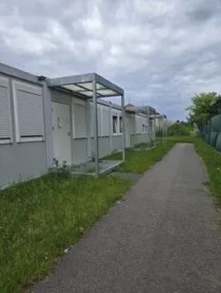 A paved path passes modular buildings with covered entrances beside a fenced grassy area.