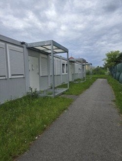A paved path passes modular buildings with covered entrances beside a fenced grassy area.