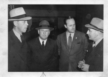 Four people in suits and hats stand together and talk indoors.