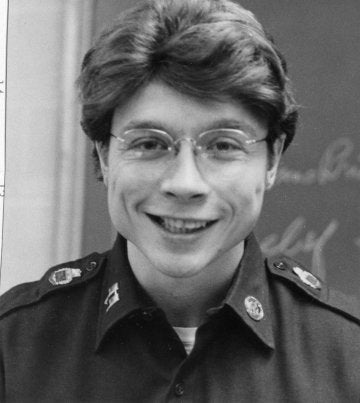 Cadet wearing glasses smiles in uniform in front of a classroom chalkboard.