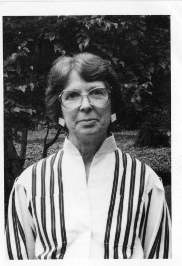 Person wearing glasses and a striped jacket stands outdoors in front of leafy trees.