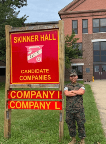 A person in camouflage uniform stands beside a Skinner Hall sign for candidate companies I and L.