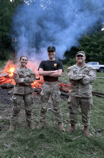 Three people stand with arms crossed in front of a large outdoor fire.