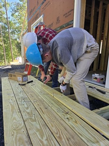 Two people in hard hats use drills to fasten wooden boards at a construction site.