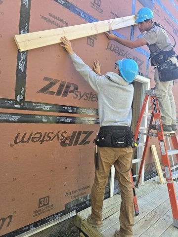 Two people in hard hats hold a wooden board against the exterior wall of a building under construction.