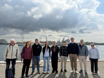 Nine people stand along a waterfront with industrial buildings and ships in the distance.