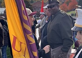 A person holding a yellow and purple flag speaks with a person in a military-style uniform at an outdoor gathering.