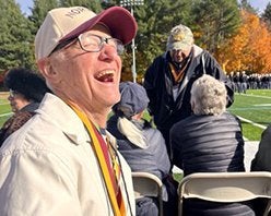 A person wearing glasses, a baseball cap, and a medal laughs while seated at an outdoor gathering near an athletic field.