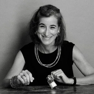 A person sits at a table holding a small jar, wearing a sleeveless top, layered necklaces, a bracelet, and a watch.