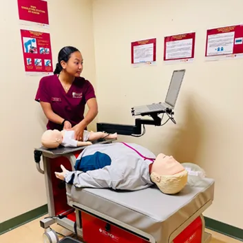 A person in scrubs practices on an infant manikin beside an adult manikin on a medical training table with a laptop on a cart.