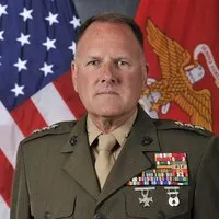 Service member in dress uniform with medals poses in front of a U.S. flag and a red military flag.