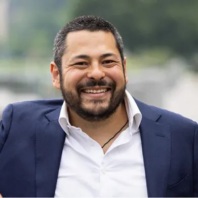 A person wearing a navy blazer and white shirt smiles outdoors.