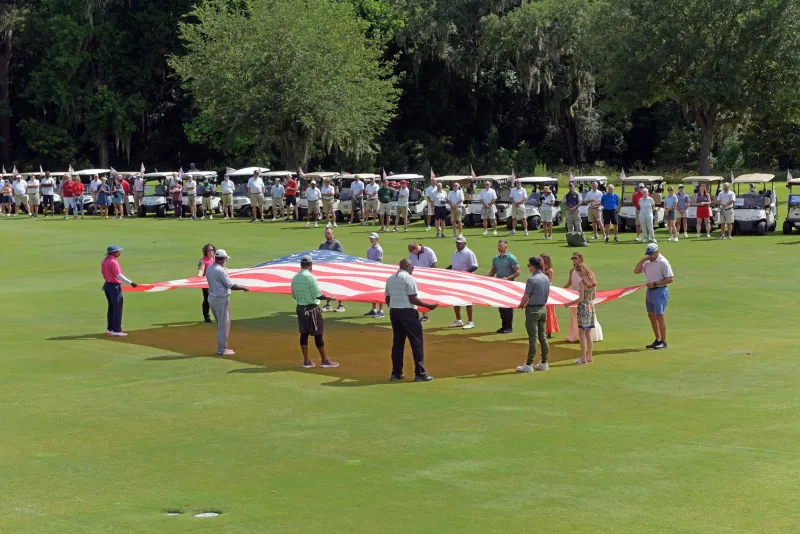 A group of people hold the American flag over a fairway green, while a line of golf carts observes from behind.