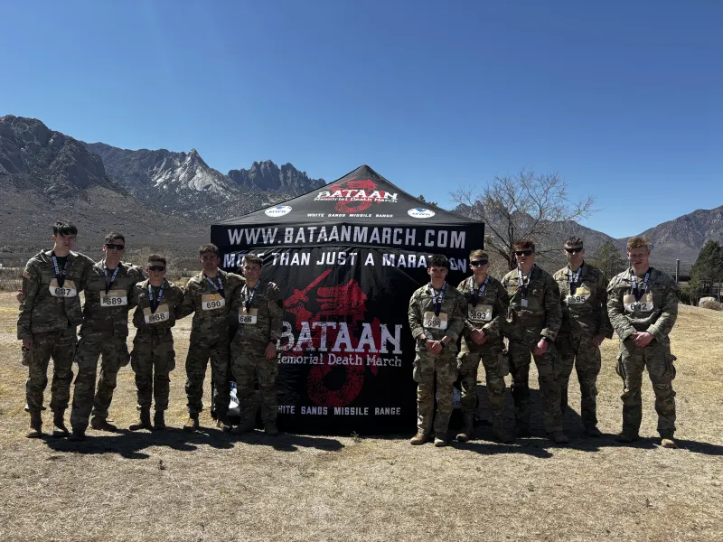 The Norwich University cadets who participated in the event stand shoulder-to-shoulder in front of a branded tent in the desert with a mountain range in the background.