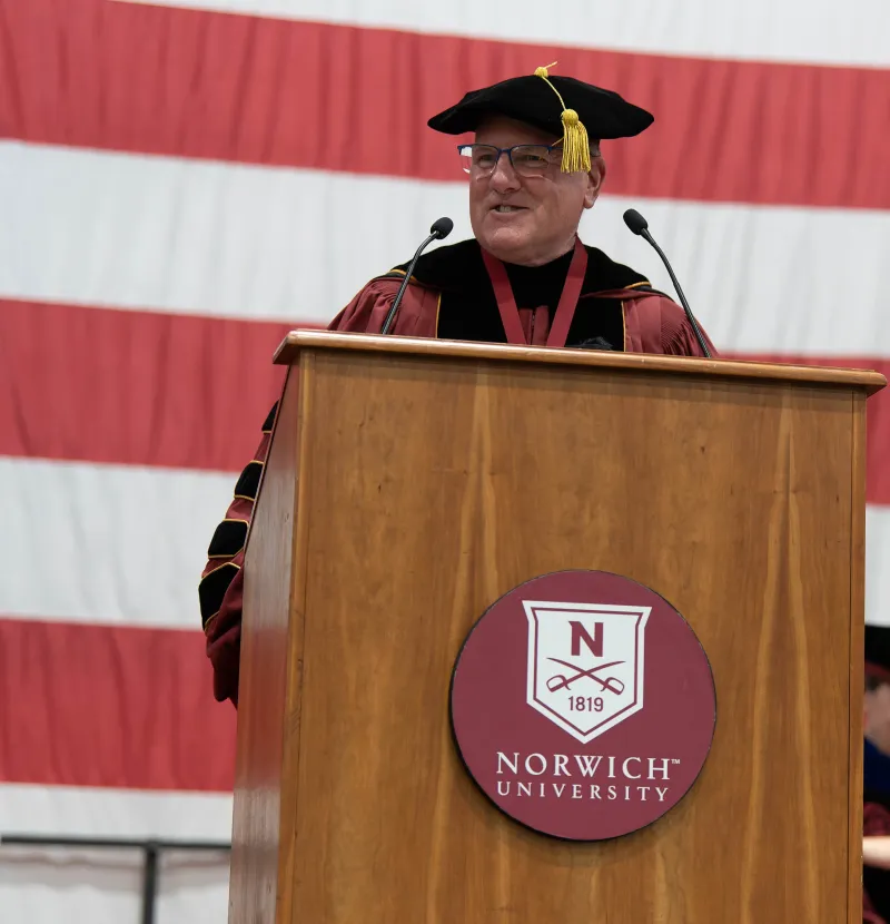 A person in academic regalia delivering a speech at a podium with the Norwich University logo, in front of a large American flag.